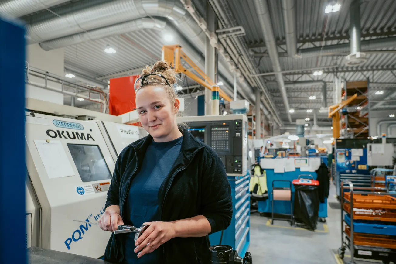 Tjej i industrimiljö håller i ett verktyg och ler mot kameran