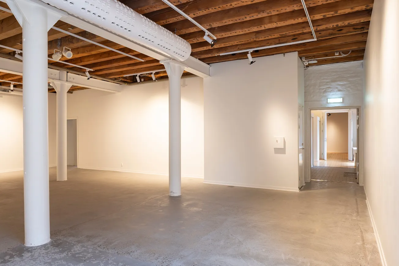 Vy över utställningslokalens ingångar, pelare, betonggolv och vita väggar.