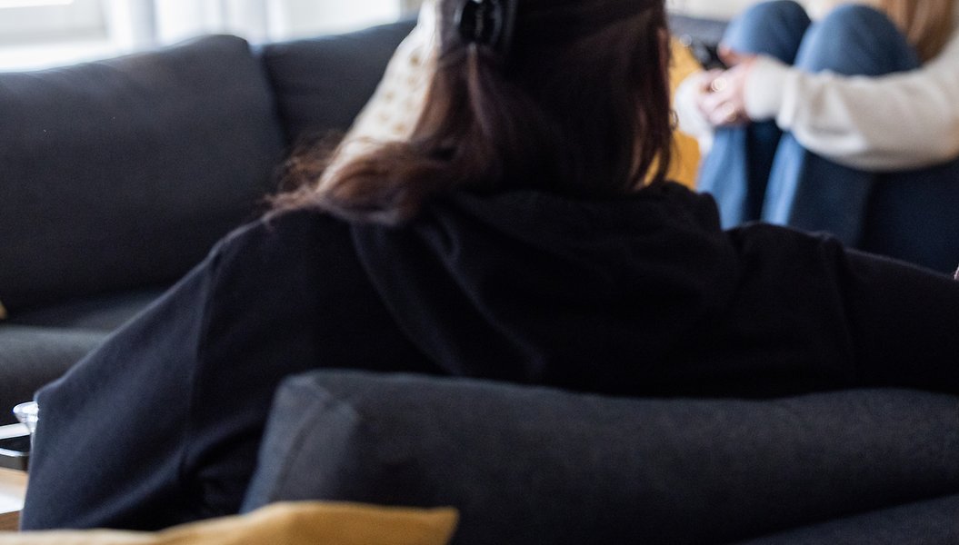 Flicka i bakgrunden som sitter i en soffa. Person med ryggtavlan mot kameran i förgrunden.