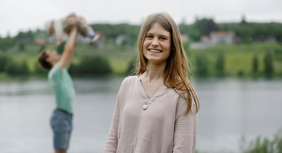 Kvinna i förgrunden som ler mot kameran, i bakgrunden skymtar en pappa med ett litet barn i famnen.
